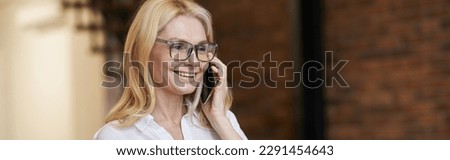 Morning. Cheerful middle aged woman with blonde hair and glasses holding a cup of coffee or tea while talking on her phone, standing at home
