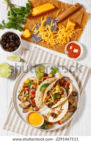 Grilled Steak Tacos with olives, tomatoes, red onion, avocado, cilantro, corn, shredded cheddar cheese and thousand island sauce on platter on white wood table with ingredients, vertical view