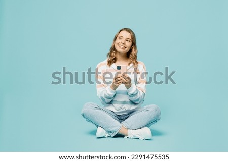 Full body minded young woman wears striped hoody sitting hold in hand use mobile cell phone look aside on area isolated on plain pastel light blue cyan background studio portrait. Lifestyle concept