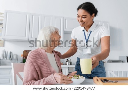 happy multiracial social worker serving lunch to smiling senior woman with grey hair Royalty-Free Stock Photo #2291498477