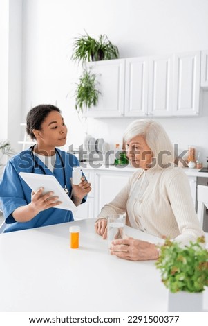brunette multiracial nurse using digital tablet and holding medication while talking to retired woman with grey hair Royalty-Free Stock Photo #2291500377