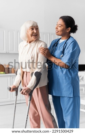 cheerful retired woman with grey hair using crutches while walking near multiracial nurse at home Royalty-Free Stock Photo #2291500429