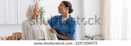 happy multiracial nurse in uniform laughing with retired woman at home, banner