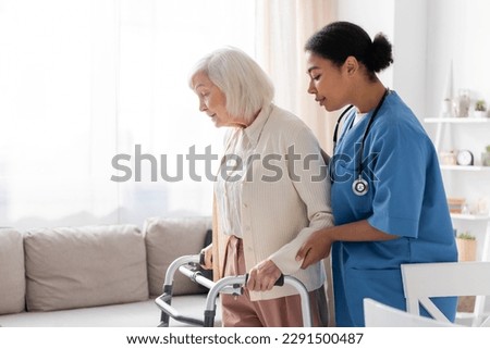 brunette multiracial nurse in uniform supporting retired woman with grey hair walking with walker at home