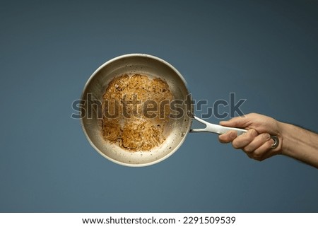 Dirty oily burnt metal frying pan held in hand by a male hand. Close up studio shot, isolated on a light blue background. Royalty-Free Stock Photo #2291509539