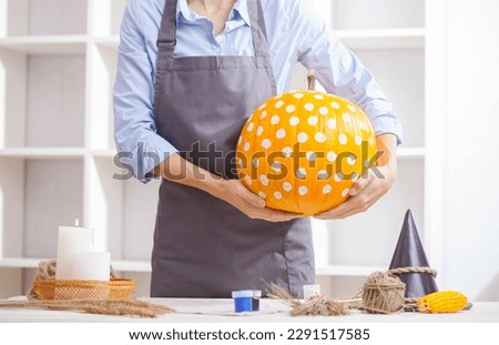 Woman in apron painting Halloween pumpkin, at wooden table, preparing holiday decorations at home