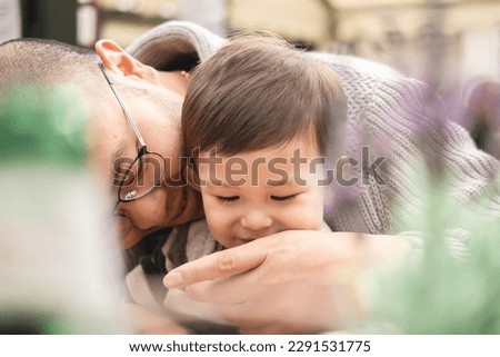 Dreamy picture of father with son, male infant sitting on his dad's legs with glasses in beer garden at pub in Edinburgh. Intimate caring moment. Bokeh with plant. Multicultural family, mixed race.