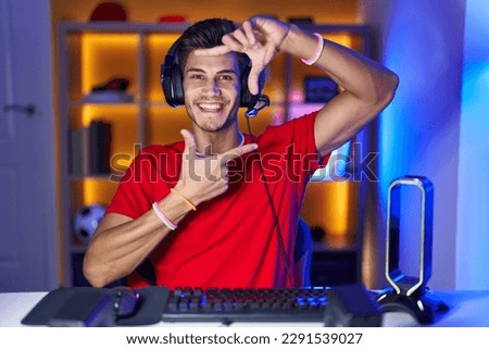 Young hispanic man playing video games smiling making frame with hands and fingers with happy face. creativity and photography concept. 