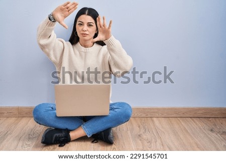 Young woman using laptop sitting on the floor at home doing frame using hands palms and fingers, camera perspective 