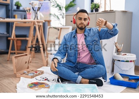 Young hispanic man painter sitting on the floor at art studio with angry face, negative sign showing dislike with thumbs down, rejection concept 