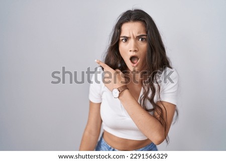 Young teenager girl standing over white background surprised pointing with finger to the side, open mouth amazed expression. 