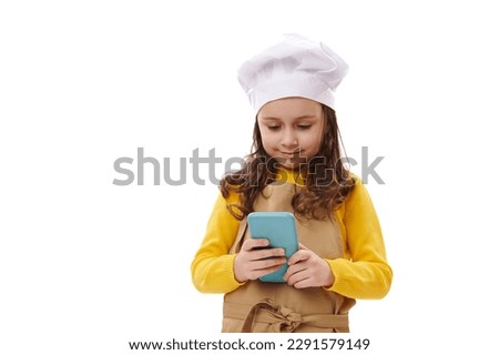 Beautiful child girl, little baker confectioner wearing white chef's hat and beige apron, checking smartphone, enyoying new mobile application. Food delivery service. Advertising shot white background