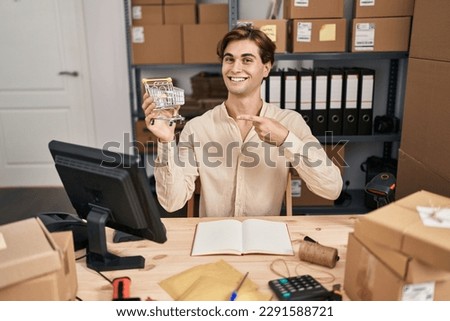 Young man working at small business ecommerce holding cart smiling happy pointing with hand and finger 