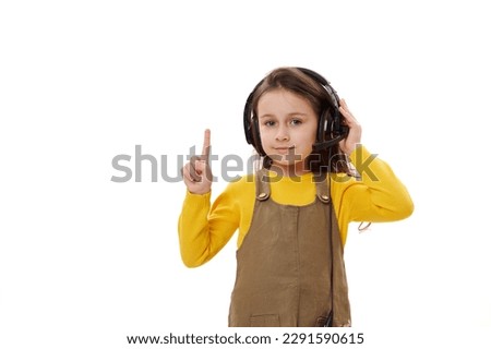 Beautiful little girl, first grader wearing headphones for online education or distance learning, pointing her index finger on a copy space, smiling looking at camera, isolated on white background. Royalty-Free Stock Photo #2291590615