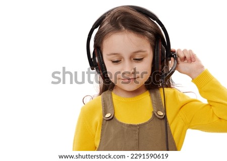 Mischievous little kid girl, primary school student, first grader with headphones, smiling over white isolated background. Smart pupil enjoys online lesson on audio headset. Distance education concept Royalty-Free Stock Photo #2291590619