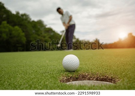 White golf ball rolling down golf hole on putting green with evening golf course backdrop and blurred golfers celebrating in winning tournament.