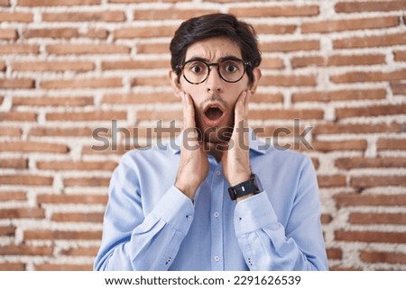Young hispanic man standing over brick wall background afraid and shocked, surprise and amazed expression with hands on face 