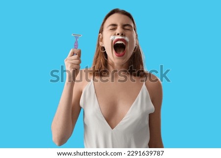 Angry young woman with shaving foam and razor on blue background