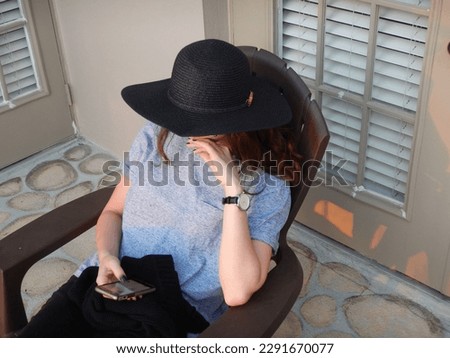 Woman wearing a hat, sitting on a deck looking at her phone Royalty-Free Stock Photo #2291670077