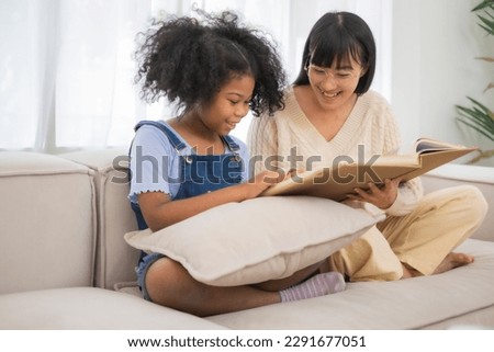 Asian mother wear eyeglass smile while teach African American daughter read the book with smile and fun at home on sofa.