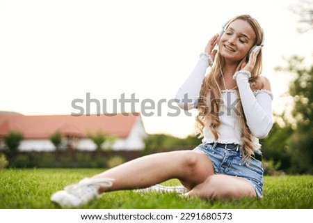 Teenager girl enjoying life and sitting in the grass in a public park, while listening to music with headphones on her head with the sunset in the background on a sunny day