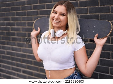 Happy young millennial student girl standing in front of a black brick wall and and looking at the camera, while holding a skateboard on her shoulder - People lifestyle concept with teenager