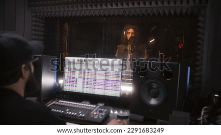 Male singer sings song in soundproof room. Audio engineer, producer uses professional DAW software with sound tracks and tools for creating music on computer. Sound recording studio. Music production.