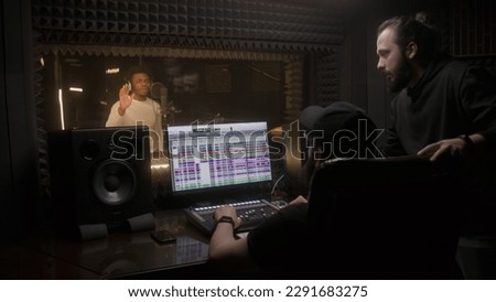 African american man records vocal in sound recording studio. Sound engineer and manager of singer look at vocalist work. Program and tools for creating music on computer monitor. Music production.