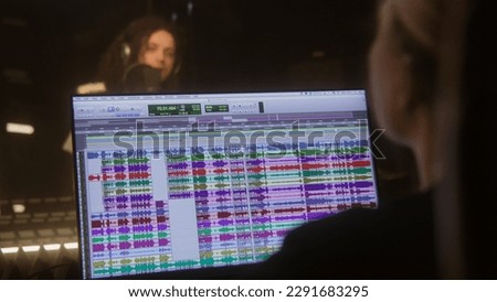 Professional singer in headphones sings lyrical composition into microphone. Sound engineer works in audio recording studio and edits song. Program and tools for creating music on computer monitor.