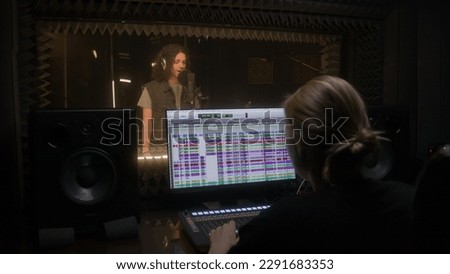 Experienced female sound engineer works with singer in audio recording studio. Man sings composition into microphone in soundproof room. Computer screen showing professional program. Music production.