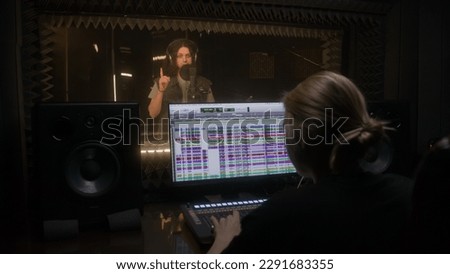 Experienced female sound engineer works with singer in audio recording studio. Man sings composition into microphone in soundproof room. Computer screen showing professional program. Music production.