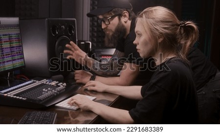 Female audio engineer uses mixing board, creates new song or DJ set with producer in sound recording studio. Computer screen showing DAW software interface with sound tracks. Music production concept.