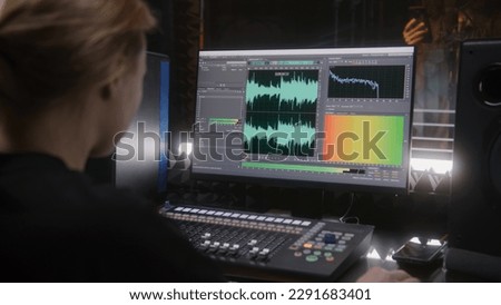 Male singer records song in soundproof room. Female audio engineer, producer uses control surface. Computer screen showing DAW software user interface with song tracks. Work in sound recording studio.