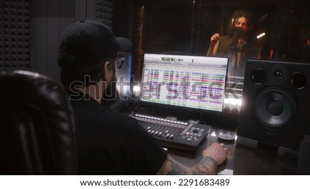 Male singer sings song in soundproof room. Audio engineer, producer uses professional DAW software with sound tracks and tools for creating music on computer. Sound recording studio. Music production.