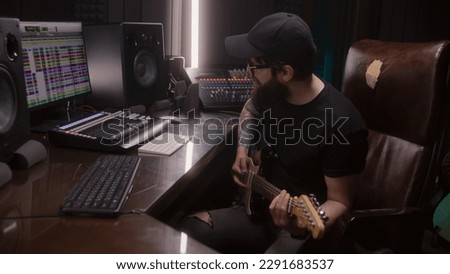 Sound engineer, musician plays electric guitar, records melody for creating new song. Computer screen showing DAW software interface with sound tracks. Professional recording studio. Music production.