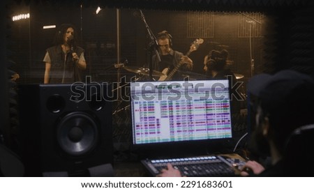 Musical band records song in recording room. Sound engineer or producer uses control surface. Computer screen showing DAW software interface with song tracks. Sound recording studio. Music production.
