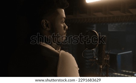 Professional male vocalist prepares to record cool track in front of microphone in soundproof room. African American singer works in sound recording studio. Concept of music production. Back view. Royalty-Free Stock Photo #2291683629