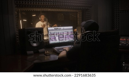 African American man in professional headphones sings new song to microphone in sound recording studio. Sound engineer records vocal by digital audio workstation software. Concept of music production.