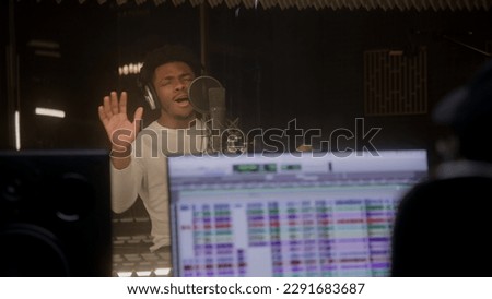 African American vocalist in professional headphones sings lyrical composition into microphone in sound recording studio. Digital audio workstation software on monitor. Concept of music production.