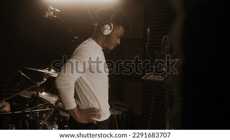 African American singer in professional headphones prepares to record new track in front of microphone in soundproof room. Vocalist works in sound recording studio. Drum set at background. Back view.