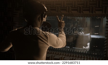 African American vocalist puts on professional headphones and sings composition into microphone in soundproof room. Audio engineer edits singer vocal on computer in sound recording studio. Back view.