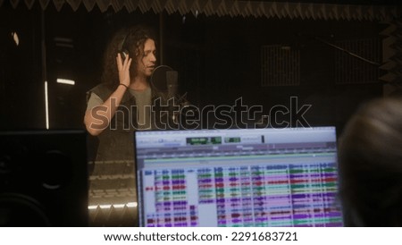 Audio engineer works with singer in sound recording studio. Vocalist in headphones sings in soundproof room. Computer screen shows DAW software interface for creating music with song playback tracks.