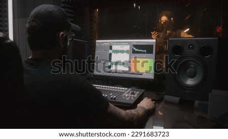 Male singer records song in soundproof room. Audio engineer, producer uses control surface. Computer screen showing DAW software interface for creating music. Sound recording studio. Music production.