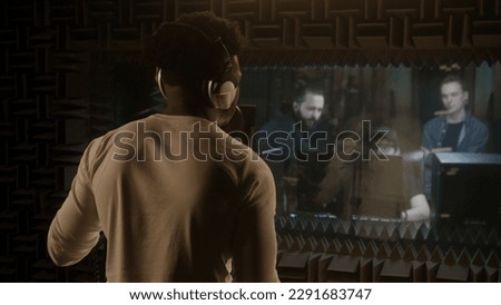 African American singer dances and sings new song into microphone in sound recording studio. Musical band and experienced audio engineer look at vocalist work. Concept of music production. Back view.