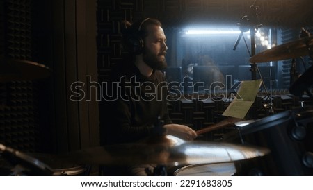 Male musician in headphones plays a drum set. Drummer records song in soundproof music recording room. Audio engineer on background. Work in the sound recording studio. Concept of music production.