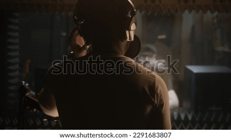 African American vocalist sings composition into microphone in soundproof room. Experienced audio engineer or producer works with singer in sound recording studio. Music production concept. Back view.