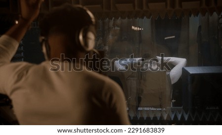 African American singer in headphones sings composition into microphone in soundproof room and gesticulates to audio engineer. Multi ethnic people work together in sound recording studio. Back view.