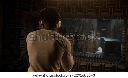 African American singer in headphones sings composition into microphone in soundproof room and gesticulates to audio engineer. Multi ethnic people work together in sound recording studio. Back view.