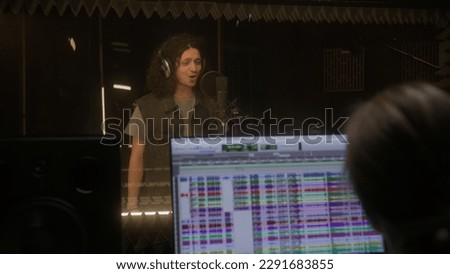 Vocalist in professional headphones sings lyrical composition into microphone in sound recording studio. Program and tools for creating music on computer monitor. Digital audio workstation software.