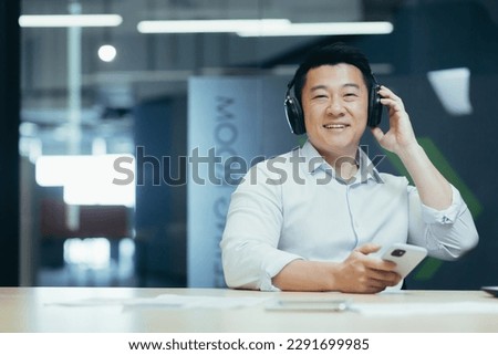 Portrait of a young Asian man sitting in the office at the table in headphones, holding the phone. Smiling at the camera.